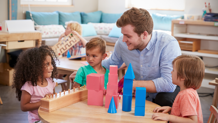 Man sitting at round table with three young children playing with coloured building blocks, one of the children is playing with a wood game with pegs in it