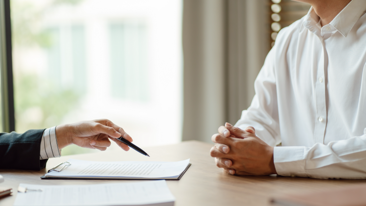 Hand holding a pen and pointing to a spot on a pad that another person sitting across the desk with folded hands is facing