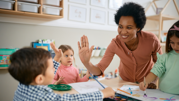 Woman at a table giving a high-five to a young boy with two other girls at the table who are painting with watercolour paints