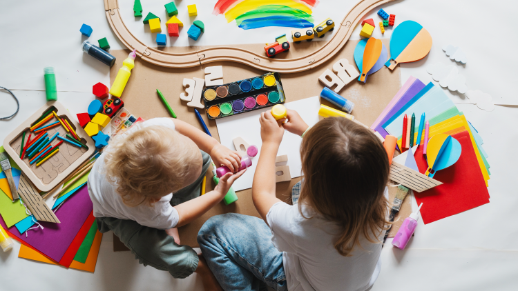 Looking down at two young children using paints from tubes on a piece of paper while surrounded by various art supplies and toys