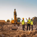 Three constructions workers stand in a pile of rubble with a dump truck and excavator in the background.