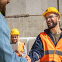 Two construction workers shake hands while a third worker looks at a tablet in the background