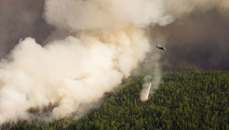 A helicopter performs a water bucket drop over a dense forest as thick smoke billows from a wildfire below. The smoke obscures much of the background, with flames hidden beneath the canopy.