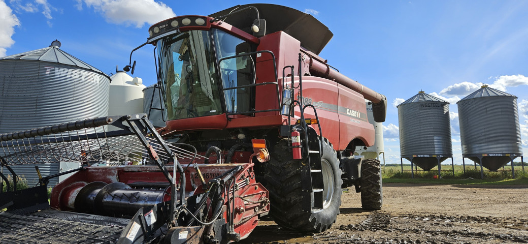A combine harvester sits in front of a row of grain bins. The sky is bright in the backdrop, with a few clouds 