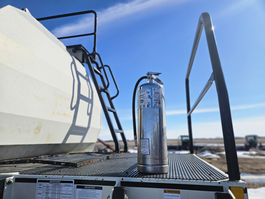 A silver fire extinguisher sits atop a piece of farm equipment. In the background the sky is blue over a farmers field. Other machinery can be seen. 