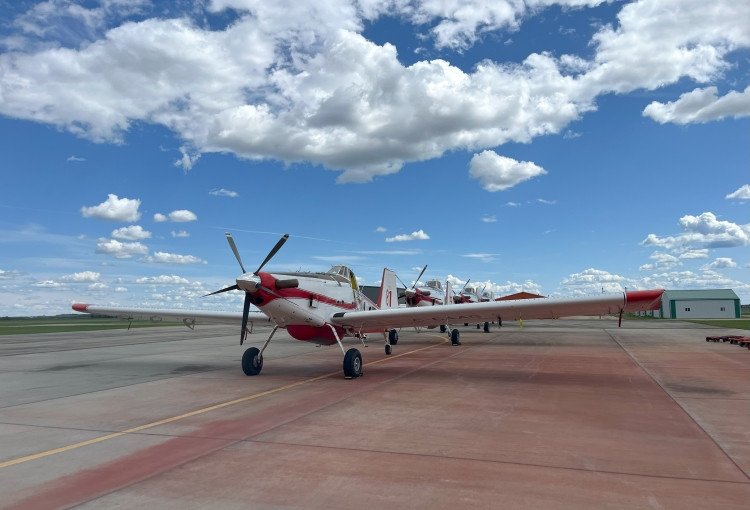 Four small propeller aircraft sit on a tarmac, lined up behind one another. The planes are primarily white with red accents, including red tips on the wings and tail. A blue sky with white clouds are in the background. 