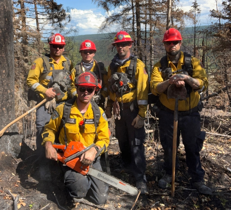 Five wildland firefighters wearing yellow shirts, red helmets, and backpacks pose in a burned forest area. One firefighter kneels in front holding a chainsaw, while the others stand behind holding firefighting tools. Burned trees and charred ground are visible around them.