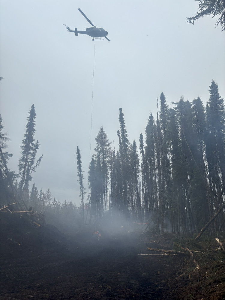 A helicopter hovers above a smoky forested area, with a orange water bucket attached to a line that sits on the ground. Below, a cleared path runs through charred ground and burned trees, with smoke rising from the forest floor. Tall trees surround the scene under an overcast sky.