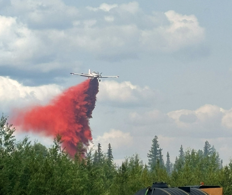 An airtanker drops a stream of red wildfire retardant over a forested area. Trees line the foreground while the plane flies just above the treetops under a partly cloudy sky.