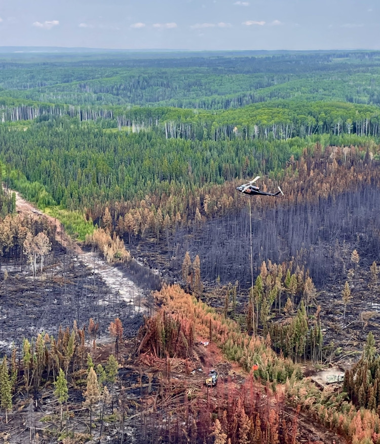 An aerial view of a wildfire burn area showing a mix of scorched and green forest. A helicopter is mid-air with a water bucket suspended beneath it, while heavy equipment works on the ground near a cleared path through the burned trees.