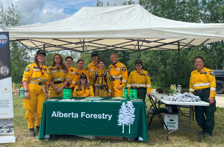 A group of Alberta Wildfire staff and Junior Forest Rangers pose for a photo under a white event tent at Saskatoon Island Provincial Park. They are standing behind an Alberta Forestry table covered with outreach materials and green water bottles. Trees and a cloudy sky are visible in the background.