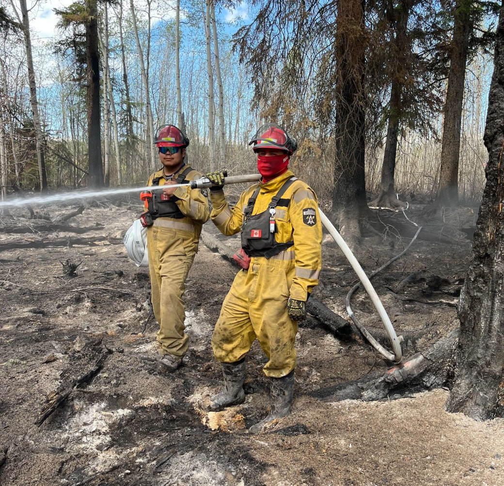 Two firefighters in yellow uniforms use a hose to spray water on a wildfire. The ground is covered in ash and trees are burnt in the background.  