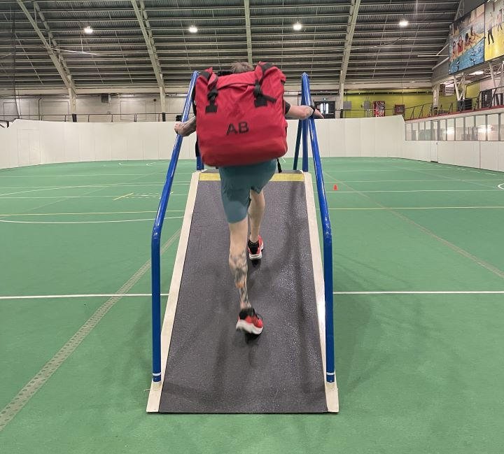 A wildland firefighter wearing a heavy red backpack is walking up a steep ramp as part of fitness testing.