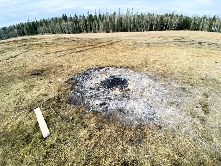 A large burnt patch of scorched grass and ash in an open field, with tire tracks visible across the dry ground and a line of leafless and evergreen trees in the distance under a cloudy sky