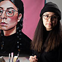 Lauren stands next to a self-portrait, holding a palette. There's a tray of paints next to her on a table.