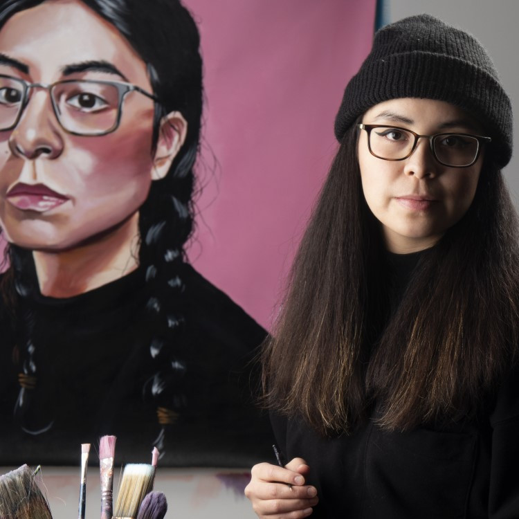 Lauren stands next to a self-portrait, holding a palette. There's a tray of paints next to her on a table.