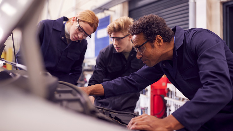Two apprentices watch an instructor work on a vehicle engine