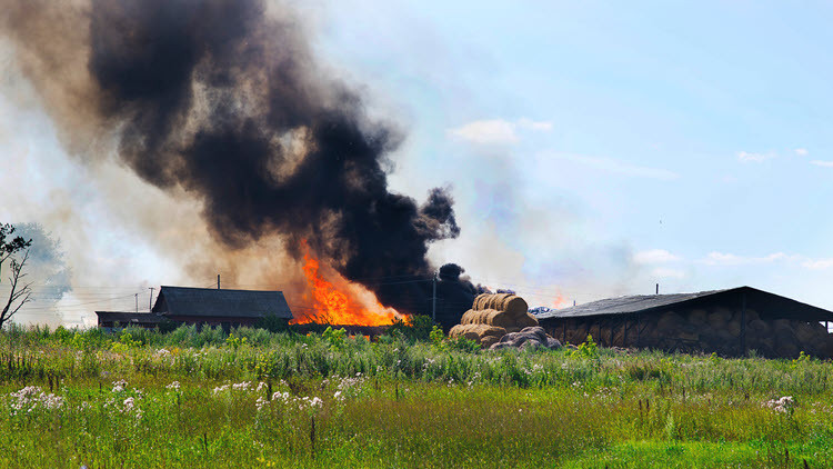 Farm in a field on fire, flames and black smoke rising in the blue sky