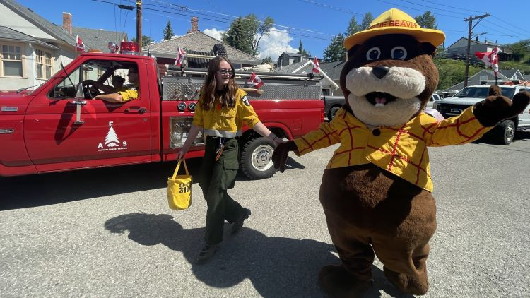 A person in a yellow and green Alberta wildfire uniform walks hand-in-hand with a large beaver mascot dressed in a yellow plaid shirt and a yellow hat labeled "BERTIE BEAVER." They are participating in a parade, walking beside a historic red fire truck decorated with Canadian flags. The scene takes place on a sunny day in a residential neighborhood with houses, trees, and a partly cloudy sky in the background.