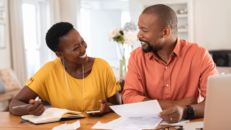 A couple sits at their kitchen table with a bunch of papers. They look at each other and smile.