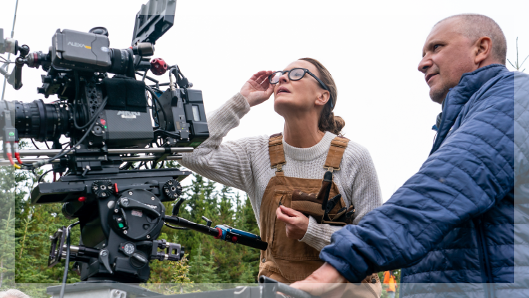 Woman wearing glasses and tan overalls and a man in a blue jacket looking at a movie cameral