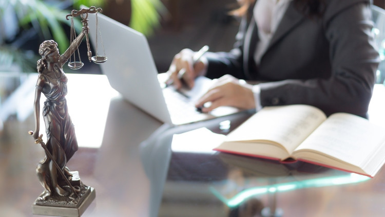 Silver lady of justice figurine holding up a two-tray scale in left hand and a sword in her right hand by her side. In the background: person in a suit working on a laptop with a book on the table