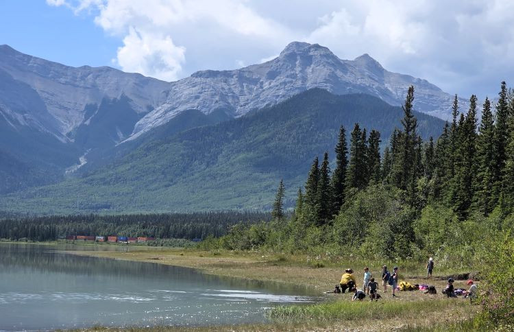 A firefighter is showing kids how to use a water pump and hose next to a river with a mountain in the background.