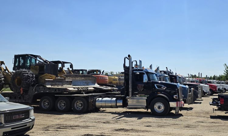 Heavy spec trucks with heavy pieces of equipment are in a row on a gravel yard.
