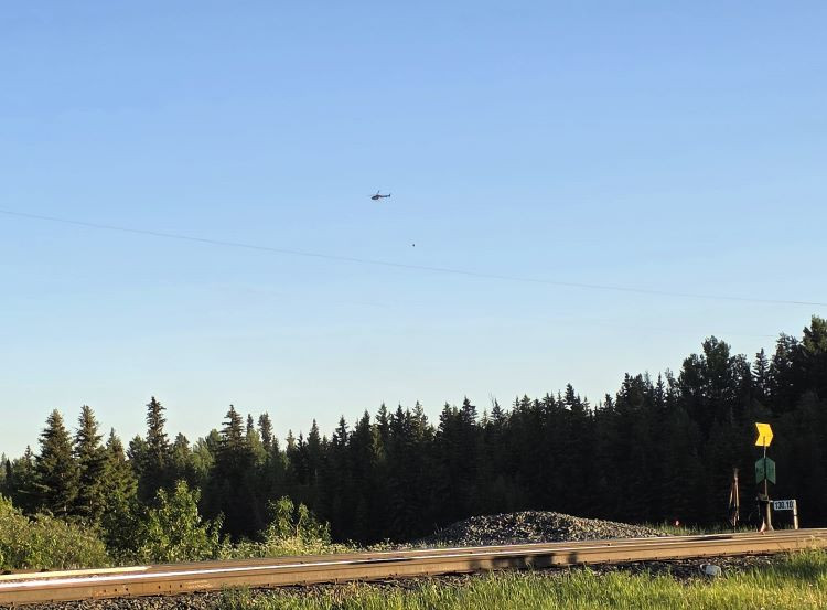 Helicopter flying over trees with a bucket. Train track at the front.