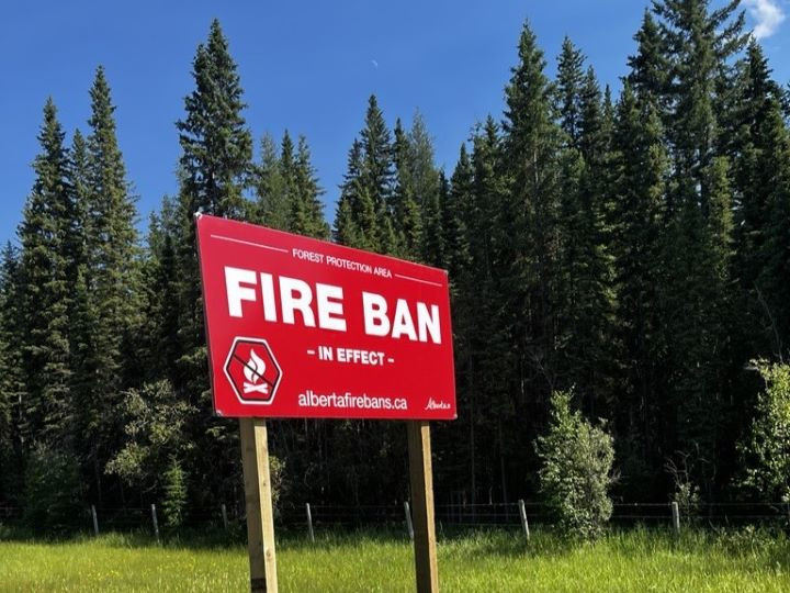 A red fire ban signs is posted in a forested area. The sign read, "Forest Protection Area Fire Ban in effect. albertaforebans.ca" and has a symbol depicted a crossed out campfire.