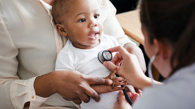 A young baby sits on their mother's lap while a doctor holds a stethoscope on their chest. The mom holds the baby's arm while the baby grabs the stethoscope cord.