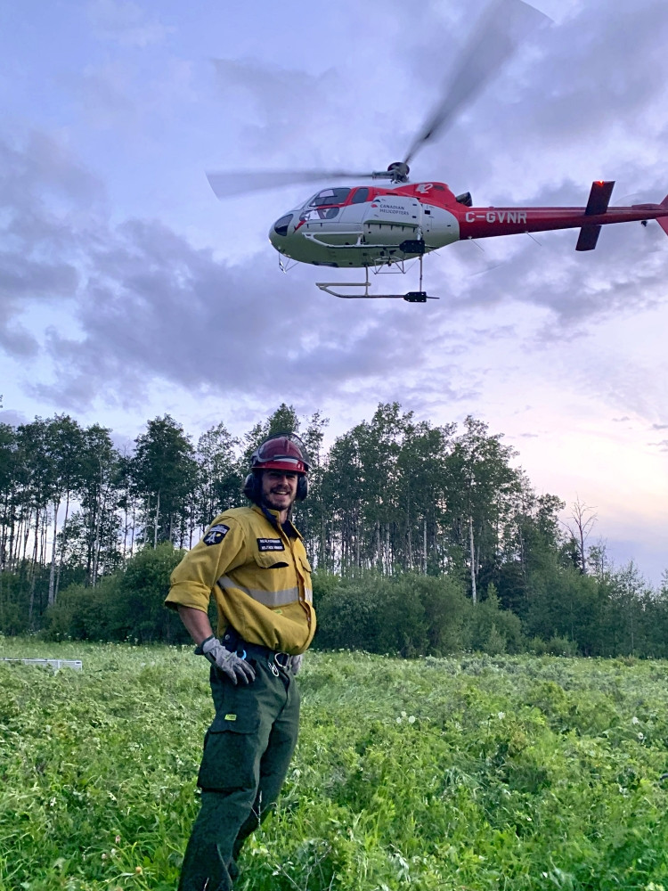 An Alberta wildland firefighter stands in a grassy clearing with a helicopter hovering in the background, with a tree line and an evening sky.