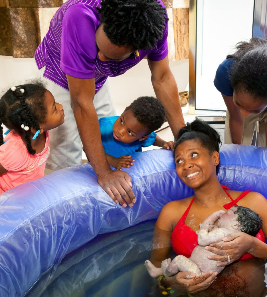 A mother in a birthing pool holds her newborn while surrounded by her smiling family.