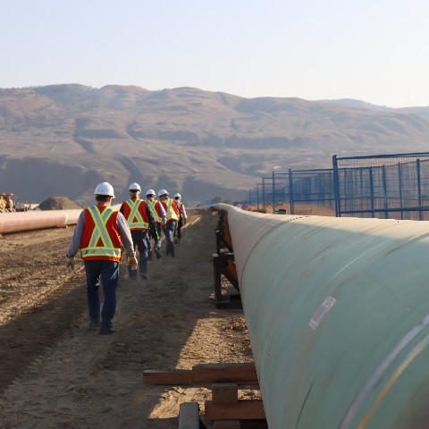 Workers in safety vests walking next to a pipeline with mountains in the background