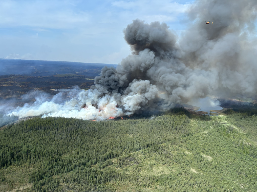 A plume of smoke rises from the trees, a yellow helicopter flies overhead