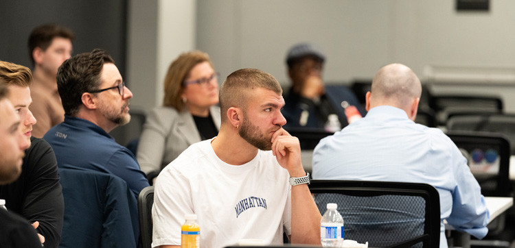 A man sits with his hand at his chin, watching a presentation, surrounded by people at a presentation  at a well-lit, indoor setting.