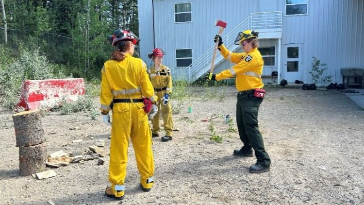 Two Junior Forest Rangers in yellow protective gear stand outdoors near a white building, watching an instructor demonstrate how to use an axe. Logs and wood pieces are scattered on the ground, and trees surround the area.