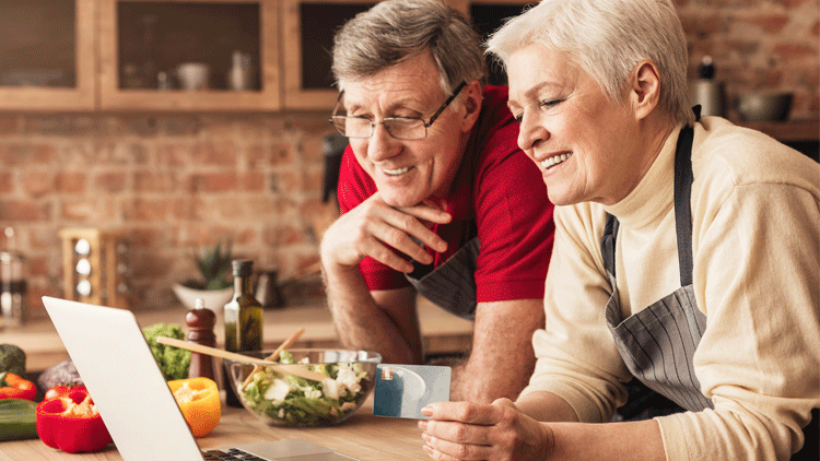 A couple lean on their kitchen counter to order take-out on their laptop. She holds a credit card and smiles. There is a salad and vegetables sitting on the counter beside them.