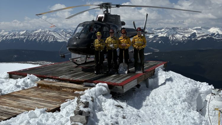 A wildland firefighter crew of four people stand in in front of a helicopter, holding snow shovels. The helicopter is parked on a wooden helipad on top of a mountain. There is snow around the helipad. There are mountains in the background horizon. 