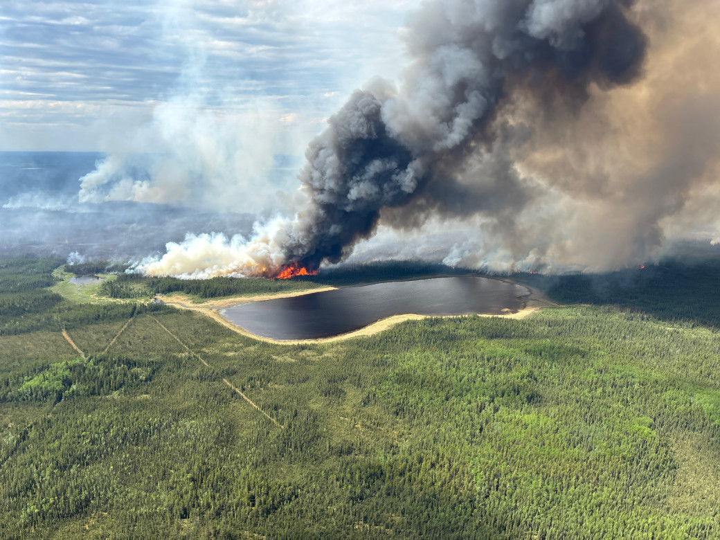 Thick smoke rises from the forest in an aerial photo. Small flames can be seen near the base, next to a small lake..