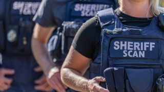 Close-up of law enforcement officers wearing tactical vests labeled ‘Sheriff SCAN.’