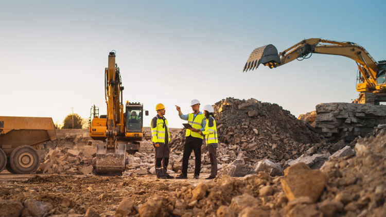 Three constructions workers stand in a pile of rubble with a dump truck and excavator in the background.