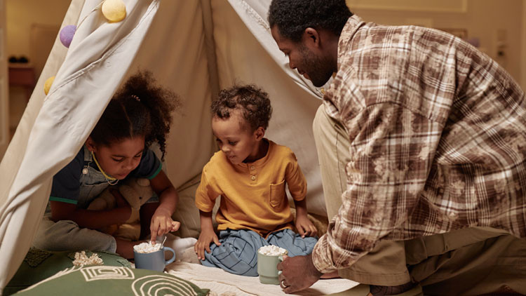 A dad plays with his two kids in a sheet tent in their living room.