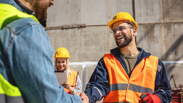 Two construction workers shake hands while a third worker looks at a tablet in the background