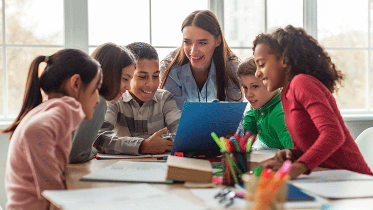 A teacher and 5 grade-school students gather around the end of a table for her to show them something. They're all smiling.