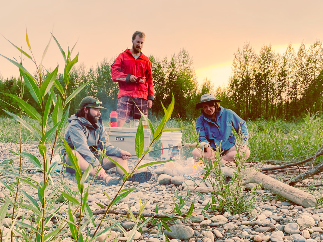 A group of people sit around a campfire at sunset. One camper roasts bannock wrapped around a stick, the other two in happy conversation