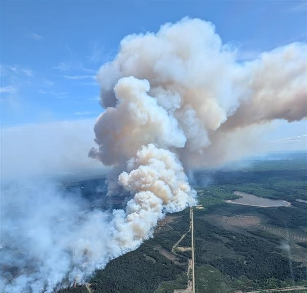 Aerial view of a large wildfire burning through a forested area. Thick white and grey smoke billows high into the sky, drifting over a grid of access roads and mixed terrain. 