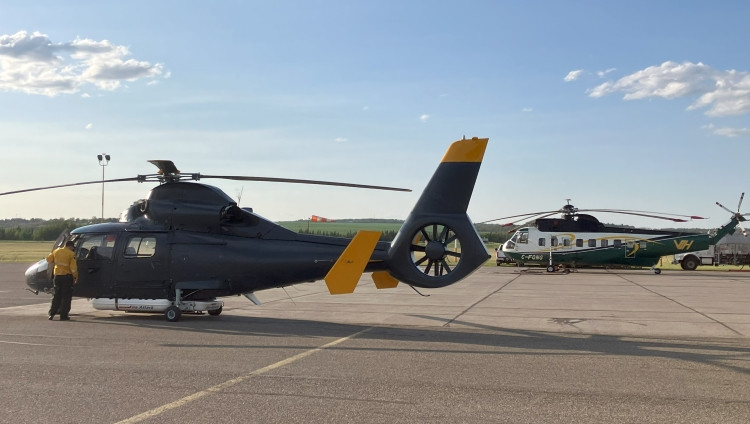 Two helicopters are parked on an airfield under a clear sky. In the foreground, a dark grey helicopter with yellow tail markings is being inspected by a firefighter in yellow gear. In the background, a larger green and white helicopter is also on the tarmac, with grassy fields and hills visible beyond the runway.