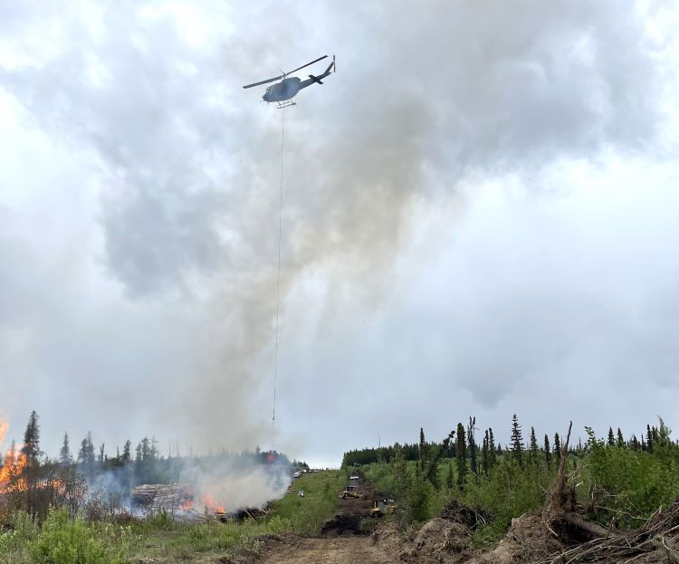 A helicopter hovers above a wildfire, carrying a suspended bucket used for aerial firefighting. Smoke rises from burning vegetation and log piles on the ground, while heavy equipment works along a cleared path through a forested area under a cloudy sky.