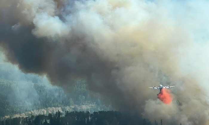 An air tanker drops red fire retardant over a dense forested area as thick smoke billows into the sky from an active wildfire.